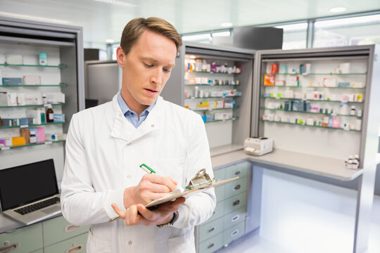 Male pharmacist wearing lab coat writing on clipboard with green pen behind pharmacy counter