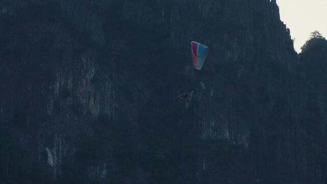 A blue paraglider is flying above the mountain in the late afternoon. 