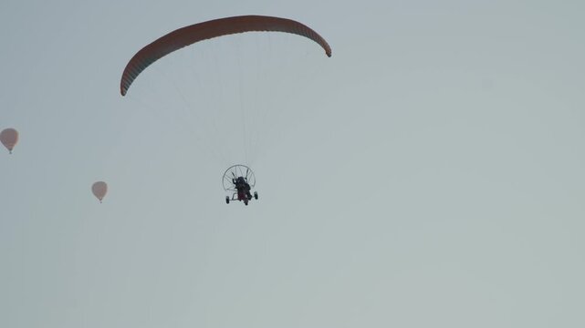 An orange paraglider is flying above the mountain in the late afternoon with a passenger.