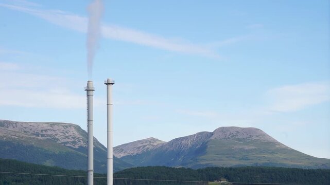 Two industrial chimneys stand in a mountainous, natural landscape, one of them belching white smoke into a clear blue sky. Concept of pollution, emissions, climate impact