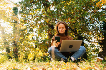 Young adult woman working outdoors in autumn park using laptop and smartphone while sitting on ground.