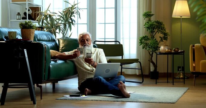 Indian mid age bearded man counting cash Indian rupees sitting on carpet in modern home, stylish mature male managing household finance and budget with paper currency indoors
