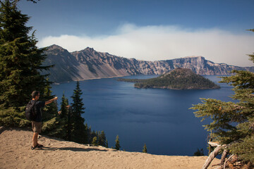 Panoramic view of Crater Lake with a cloud of smoke behind and a man pointing towards the lake in Crater Lake National Park © Conchi Martinez