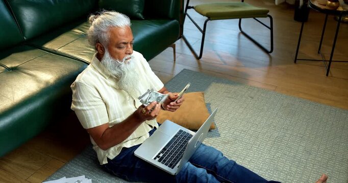 Indian mid age bearded man counting cash Indian rupees and relaxing between work sitting on carpet in modern home, stylish mature male managing household finance and budget indoors