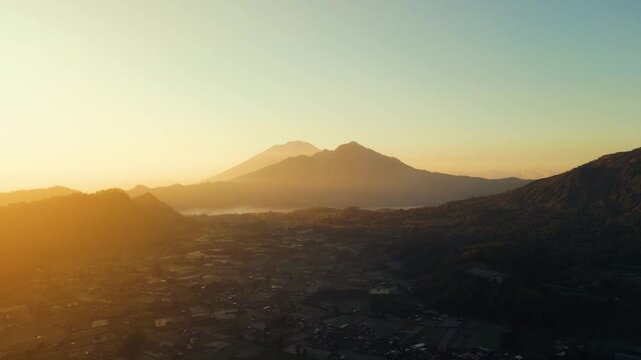 A stunning sunrise over Mount Batur and Mount Agung in Bali reveals mist-covered settlements and lush green plantations. The early light casts a serene glow over the breathtaking landscape.