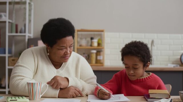 Grandparent looking at her grandson while he solving math problem, writing on paper