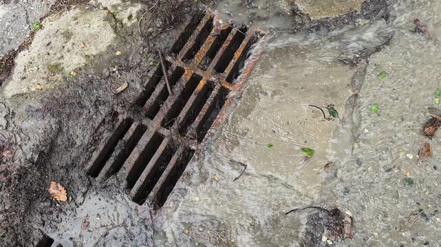 Rainwater flowing into a rusty metal sewer grate on a concrete pavement during a heavy rainstorm, draining away surface runoff in a city street environment
