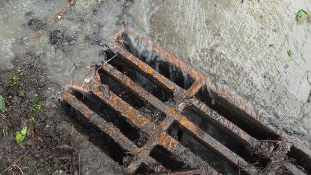 Heavy rainfall water runoff flowing into a rusty metal storm drain grate on a city street, showcasing urban drainage infrastructure during a downpour and flood prevention