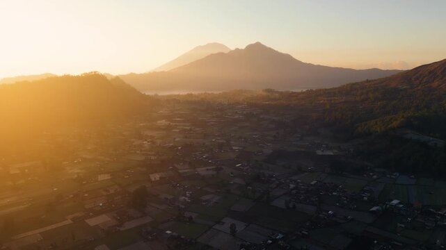 A stunning sunrise over Mount Batur and Mount Agung in Bali reveals mist-covered settlements and lush green plantations. The early light casts a serene glow over the breathtaking landscape.