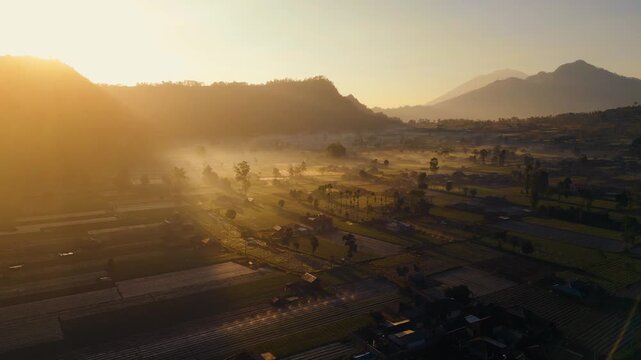 A stunning sunrise over Mount Batur and Mount Agung in Bali reveals mist-covered settlements and lush green plantations. The early light casts a serene glow over the breathtaking landscape.