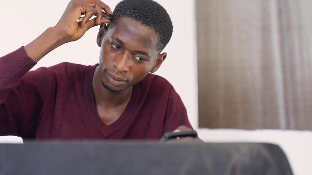 Black man painting at table, examining sketchbook and adjusting ear, thoughtful expression, indoor studio light, burgundy sweater, phone reference on table, focused pencil and brush study, gradual