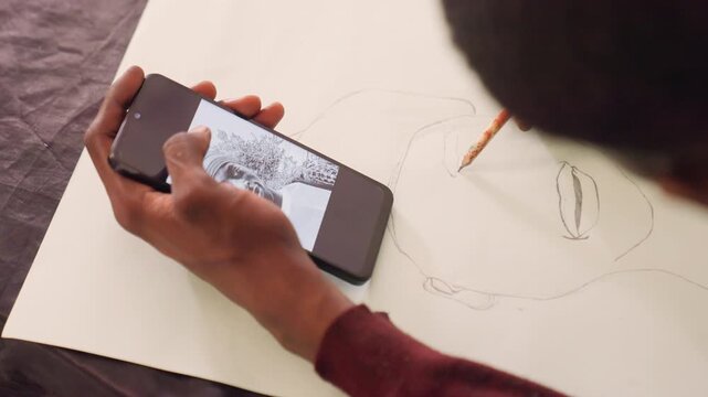 Black man composing portrait using phone reference. Topdown framing shows phone image beside pencil outlines on paper, deliberate contouring of eyes and mouth, late session creativity, raw process