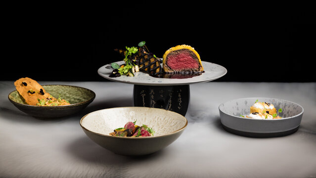 A mix of gourmet fusion fine dining dishes, including beef wellington, steak tartare, scallops and a mango cake dessert with dry ice creating a mist or smoke like effect