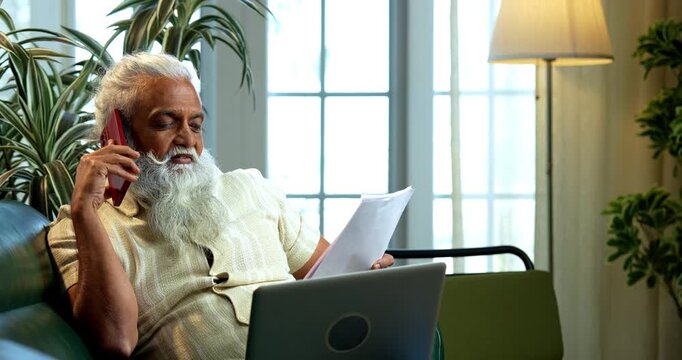 Indian mid age bearded man working online using laptop, smartphone call and reports sitting on sofa in modern home, stylish mature male multitasking business discussion and paperwork indoors