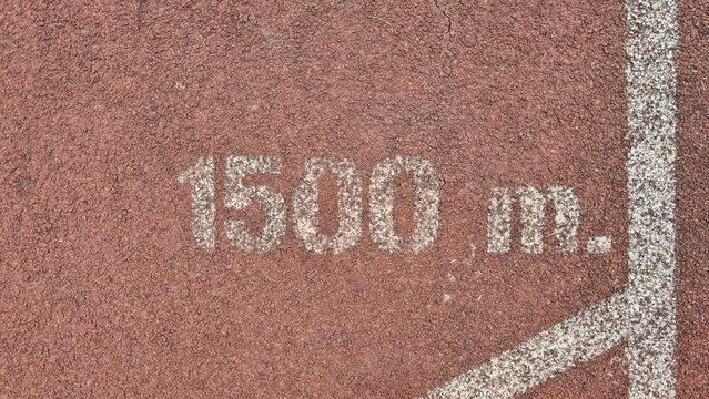 1500 m marking on red synthetic running track surface. Top view of a 1500-meter distance marker on a red rubberized track. Close-up of durable red track material with white 1500 m lettering.
