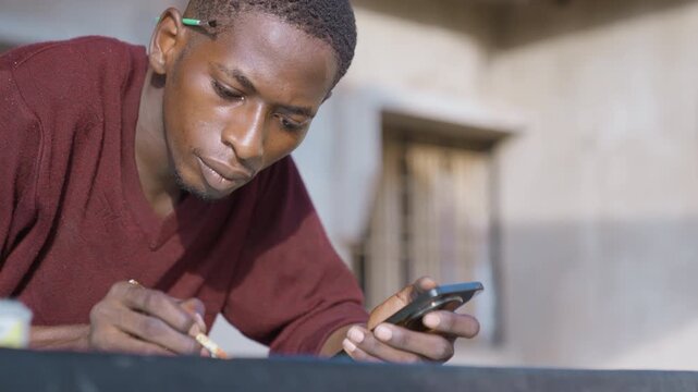 Black man painting with phone reference, focused expression, pencil tucked behind ear, indoor studio setting, tiny brush applying fine strokes on model surface, warm soft light, concrete wall
