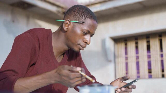 Black painting reference study, contemplative artist balancing brush and phone while checking image, maroon sweater, courtyard setting, calm focused expression and steady hand movements