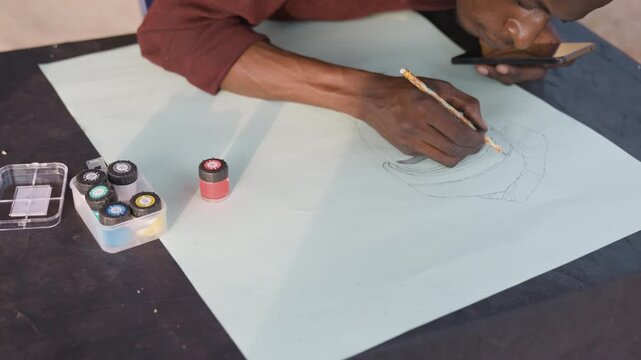 Hands drawing dotted design on paper, studio scene with pale sheet on black mat, small jars of pigment and ink, pencil tracing circular pattern, focused maker shaping mandalalike motif, quiet