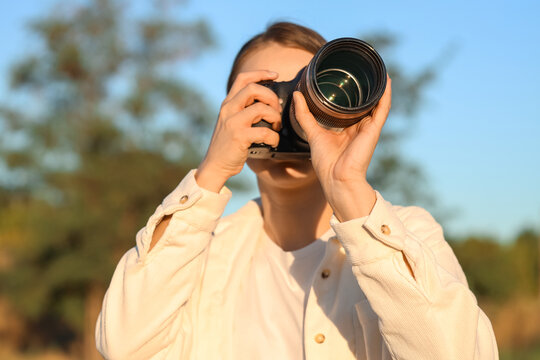 Young woman photographer holding professional camera with telephoto lens outdoors in golden hour light, close up portrait of female taking pictures in nature with blurred background