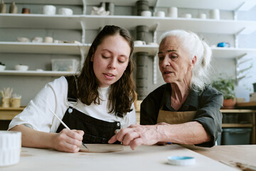 Obraz premium Caucasian young adult woman painting ceramic plate while senior Caucasian woman guiding her, both sitting at table in pottery studio, shelves with pottery visible in background