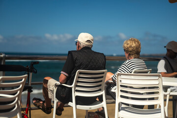Fototapeta premium Senior couple seating by a seaside promenade railing, looking out at the ocean on a sunny day