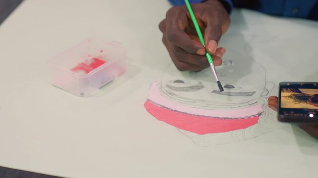Black artist painting portrait with pink highlights, closeup hand applying watercolor on canvas, phone reference showing face, plastic paint tray, demonstrative tutorial mood, focused technique