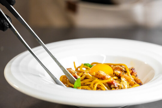 A chef meticulously plates spaghetti carbonara in a white bowl, using kitchen tongs to position the pasta near a bright egg yolk and savory guanciale