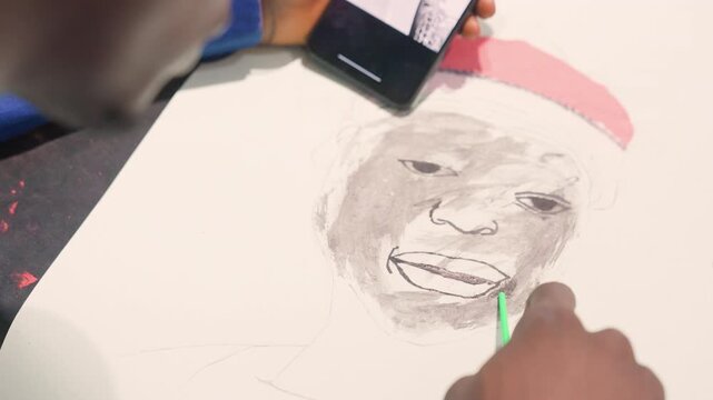 Black artist sketching portrait with phone reference. Creating black painting tonal study on paper hand guides pencil and charcoal adding shading to face and red hat close studio setting with focused