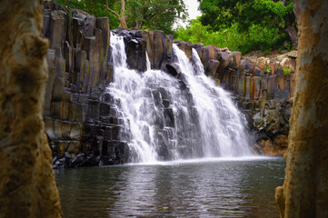 Fototapeta premium Rochester Falls in Mauritius flowing over distinctive basalt rock columns into a calm pool, surrounded by dense tropical forest and lush greenery.