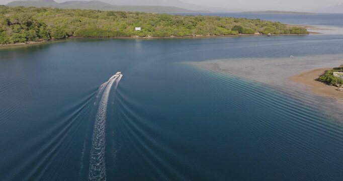 Aerial drone shot following a small boat leaving the calm lagoon of West Bali National Park, Indonesia, with rippling wake patterns spreading across deep blue tropical waters near mangrove coastline
