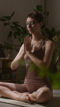 Full vertical shot of young gen Z Caucasian female yoga devotee sitting in lotus posture on mat, folding hands in namaste mudra for daily meditation, green potted plants in foreground