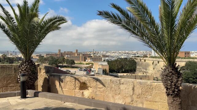 High angle panoramic view of a Mediterranean cityscape featuring modern architecture, lush green trees, and historic stone bastions seen from a terrace with palms. Malta.
