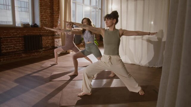 Full shot of three experienced young Caucasian women practicing yoga in loft red brick studio, performing sequence of warrior, triangle and reverse warrior asanas together in unison