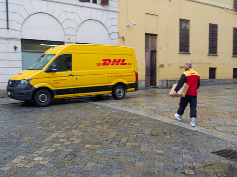 A uniformed DHL courier walks with a parcel towards a building, with a branded yellow delivery van parked nearby on a wet, historic cobblestone street