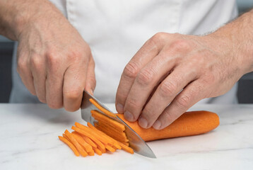 Close-up of chef hands slicing fresh carrot into julienne strips with a sharp knife on a white marble surface, food preparation technique