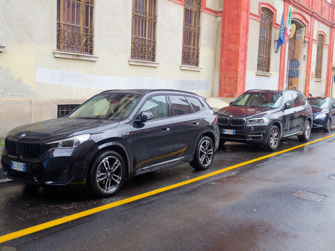 A row of modern BMW SUVs, including an X1 and an X5, are parked on a wet asphalt road with yellow lane markings, reflecting the overcast sky and surrounding architecture