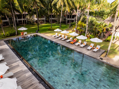 Aerial view of a long, dark turquoise pool reflecting palm trees, amidst sunbeds and white umbrellas, contrasting with the bright green grass, Prince Maurice, Flacq District, Mauritius.