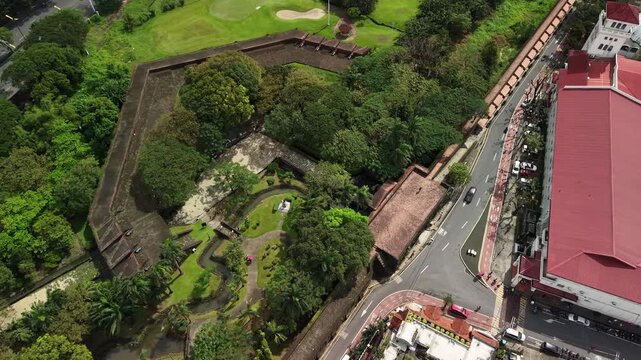 Top-down drone view of Intramuros stone ramparts and a landscaped garden with ponds beside a busy Manila street, with golf course greens and trees filling the frame in daylight.