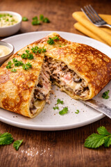 A delicious pancake stuffed with chicken, mushrooms, and melted cheese, served on a white plate with herbs.  