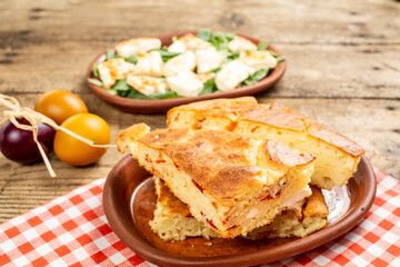Pieces of homemade bread with onions served in an earthenware bowl on a wooden table, with salad and fresh vegetables on the side.