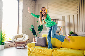 Naklejka premium Young woman enjoying music while singing on microphone at home, wearing headphones, and having fun in cozy interior