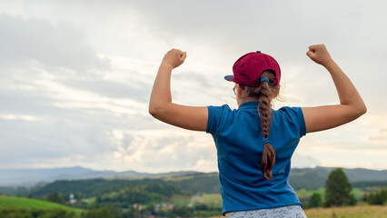 Young girl hiker with raised arms in victory pose overlooking scenic green valley hills and dramatic cloudy sunset sky