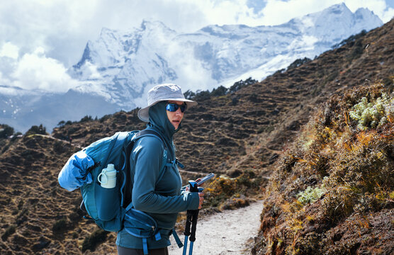 Solo female trekker with backpack checking smartphone route on mountain trail with snowy Himalayas range background during Everest Base Camp trek