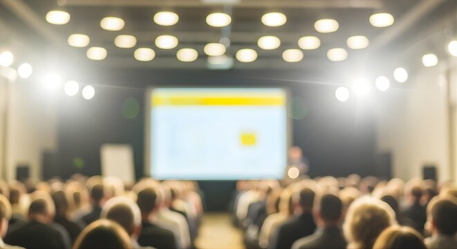 Blurred image capturing the essence of a large conference or seminar event with an audience focused on a bright projection screen and a speaker presenting to the attendees.