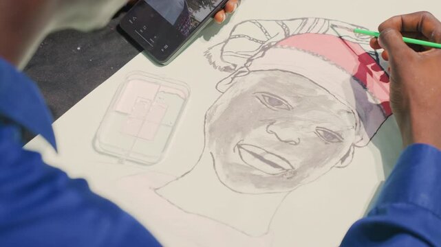 Black freelance illustrator drawing portrait painting at workshop table, steady hand fills pencil strokes into joyful black man face, smartphone reference visible, commercial commission vibe, bright