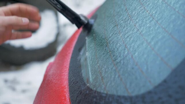 Mans hand lifts and lowers rear windshield wiper on glazed back window of red car, checking its function and clearing ice
