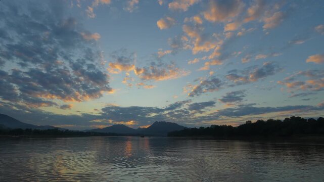 View of the sunset behind the mountais at the Mekong River with light cloudy sky and calm water in the foreground, with mountain silouettes in the background.