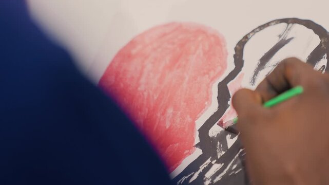 Tight frame hand outlining black painting, green brush tracing bold contour of red heart, shallow depth of field highlights textured pigment and deliberate line work, concentrated studio atmosphere