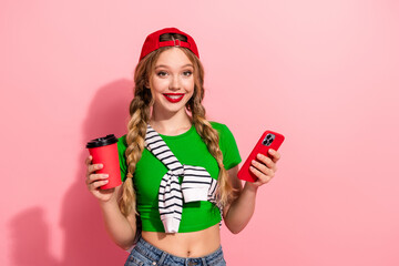Young stylish girl with green top braids red cap holding phone and coffee against pink background