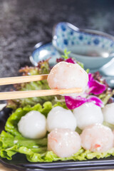 A close-up of a fish ball held by chopsticks above a plate of fish balls arranged on lettuce leaves with a purple flower garnish.
